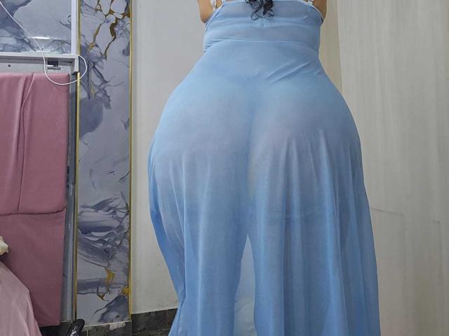 NaomyHott69's butt in a sheer blue dress, showing her ass through the fabric, standing with her back to the camera, black curly hair visible.