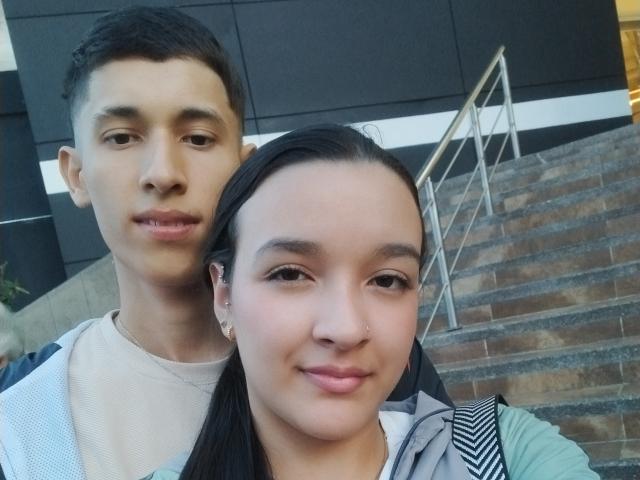 Alanna e James, com um piercing fofo no nariz, sorrindo, com cabelo escuro, em pé atrás de James, que tem cabelo preto curto e uma expressão neutra, vestindo uma camisa bege..