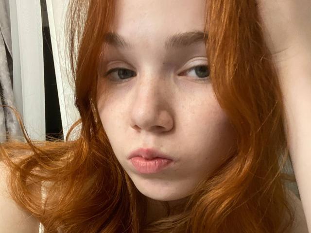 KatiRokisky has red hair, pale skin, and freckles with pouty pink lips, looking directly at the camera with a slightly bored expression. Her blue eyes are half-open.