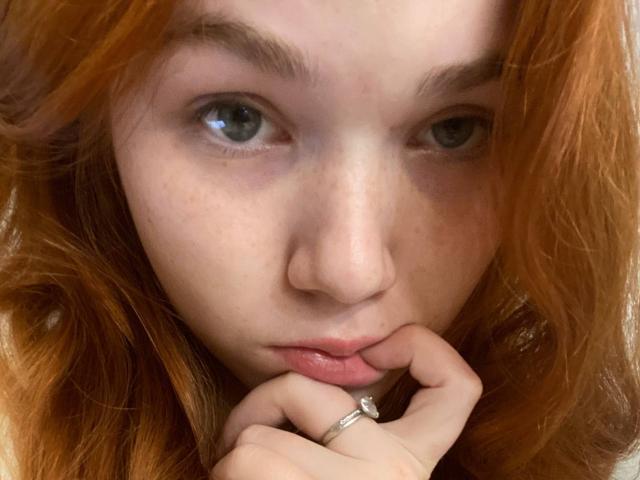 KatiRokisky has red hair, grey eyes, and freckles, with her finger on her lower lip, wearing a ring on her right hand. Her expression is serious and focused.