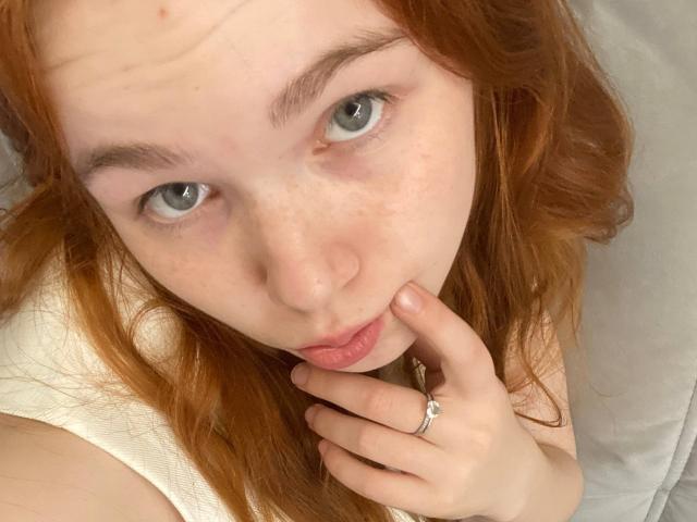 KatiRokisky has red hair, green eyes, and freckles, pouting with her finger on her cheek, wearing a white shirt, and a silver ring on her finger.