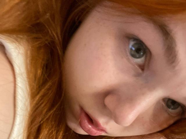 KatiRokisky's face with red hair, blue eyes, and slightly parted pink lips, lying down, showing a soft, fair-skinned, freckled face. Her expression is gentle and curious.