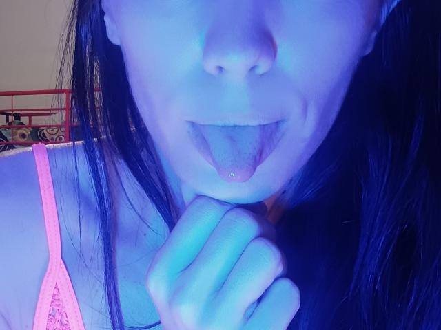 OrianaLaFrancaise's face in blue light, tongue out, hand near mouth, wearing pink top, black hair, slightly visible nose, and a playful, teasing expression.