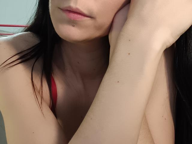 OrianaLaFrancaise's face, slightly smiling, with black hair, pale skin, red bra strap, and her arm resting on her cheek, showing a few small moles on her arm.
