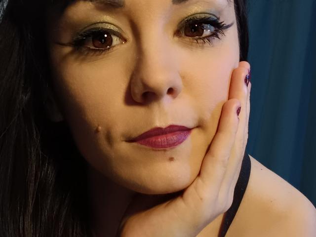 OrianaLaFrancaise's face, showing brown eyes with green eyeshadow, dark lipstick, and a small mole under her right eye. She's touching her cheek with her hand, black nails.
