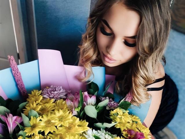 Niferttiti with curly blonde hair, thick black eyeliner, and pink lips, looking down at a colorful bouquet of yellow, purple, and pink flowers. She's wearing a black strap dress.