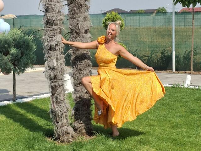 Merlynon, with yellow dress and flower in hair, stands by palm tree, knee raised, holding dress, showing off her ass, grassy yard, sunny, smiling.