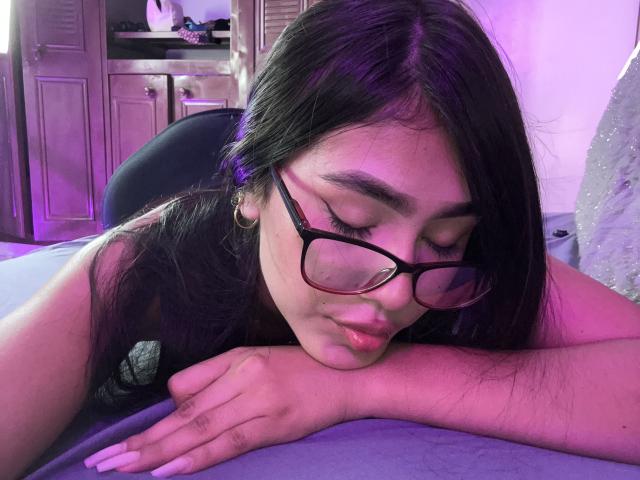 DulceNoir, with black glasses and long dark hair, rests her head and arm on a bed, pouting slightly, in purple-lit room. Her ass is partially visible.