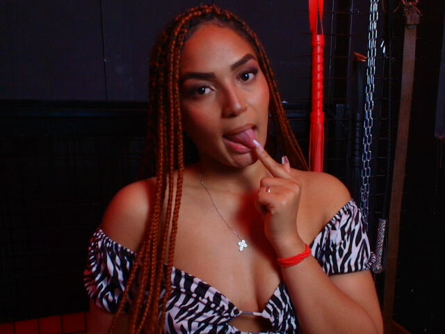 BrianaPratt with long braids, tongue out, licking finger, zebra-patterned top, showing cleavage, wearing a necklace, red bracelet, dark background, sexy, seductive.