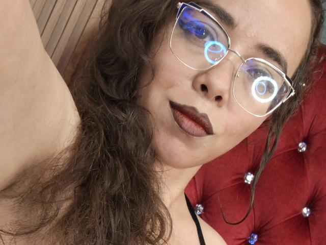BarbaraLeythonn with wavy brown hair, wearing clear glasses with blue light reflections, dark lipstick, and black strappy top, showing a neutral facial expression. Red tufted background.
