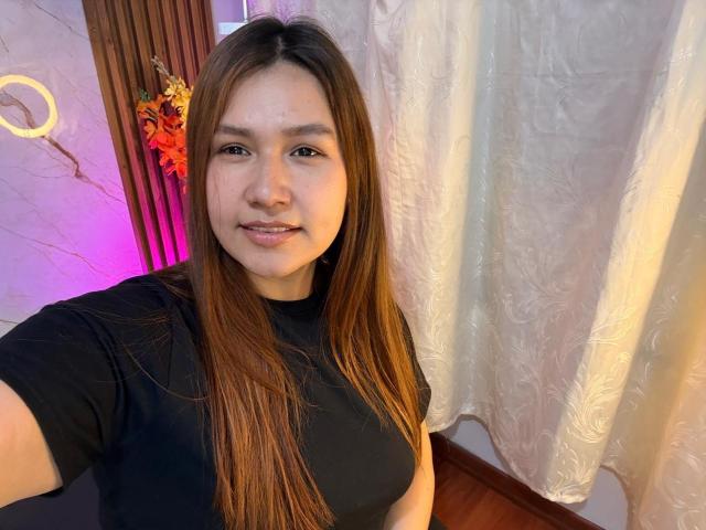 SofiaVelvet with long brown hair, black shirt, and small tits, smiling in a selfie, standing in front of white curtains and wooden wall. Her skin is light brown.
