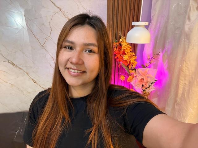 SofiaVelvet with long brown hair, black shirt, smiling, holding phone, medium tits, cute, Asian, marble wall, white lamp, orange flowers, purple light, wooden panel, selfie.