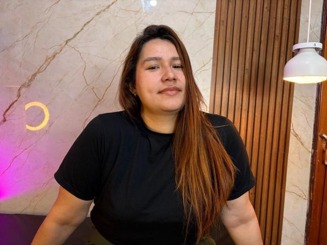 SofiaVelvet, chubby woman with long brown hair, black shirt, slight smile, standing in front of wooden panel and marble wall, under white hanging light.