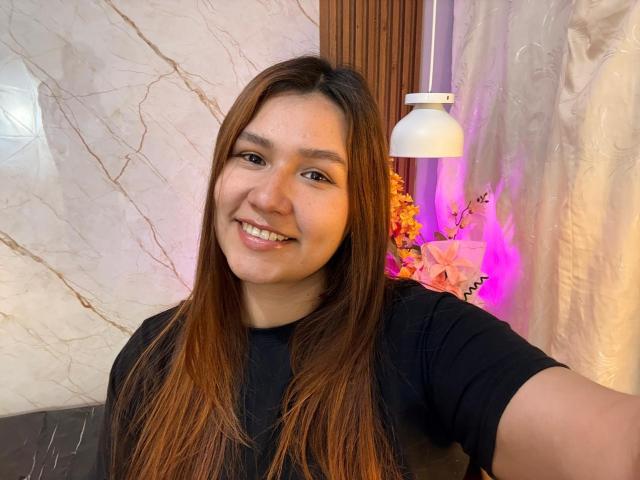 SofiaVelvet, con lunghi capelli castani, sorridente, indossa una camicia nera, ha una corporatura media, pelle chiara, occhi dolci e un trucco naturale, ed è in piedi accanto a una lampada bianca e fiori rosa..