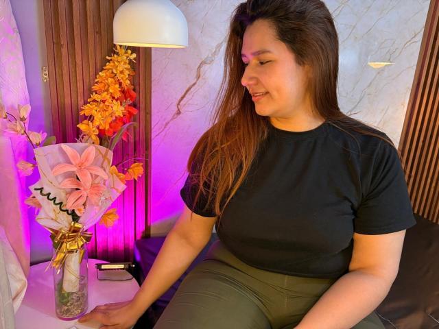 SofiaVelvet with brown hair, black t-shirt, and green pants, sitting, smiling, hands on a table with a pink flower vase and orange flowers, under purple and white lights.