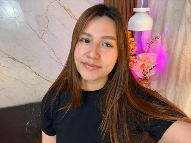 SofiaVelvet has long brown hair, a black t-shirt, and a cute smile. Her brown eyes and light skin are visible. She's standing with a white lamp behind her.