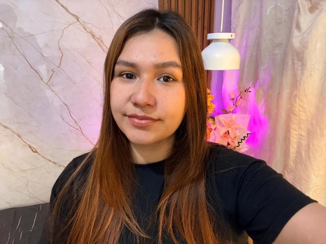 SofiaVelvet, avec ses longs cheveux bruns, son t-shirt noir et un sourire discret, se tient devant un mur en marbre blanc, avec une lumière suspendue. Ses fesses ne sont pas visibles..