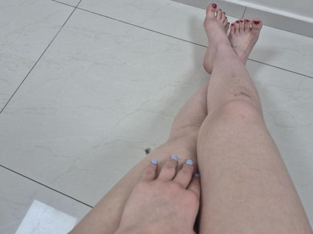 JoannaSia's pale legs with slight redness, crossed at the ankles, showing red-painted toenails and blue-painted fingernails, sitting on white marble-tiled floor.