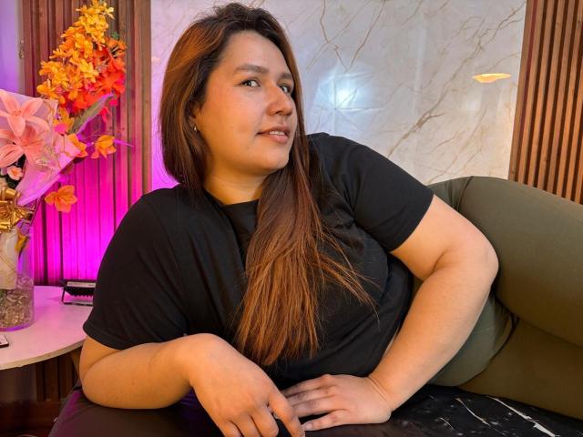 SofiaVelvet, chubby with long brown hair, wearing a black t-shirt, sitting on a green couch, looking to the side, with a pink and orange flower vase to the left.