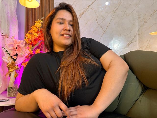 SofiaVelvet, chubby with long brown hair, wearing black tee and green pants, sitting relaxed, smiling, hands on lap, lamp and flowers in background, soft purple and yellow light.