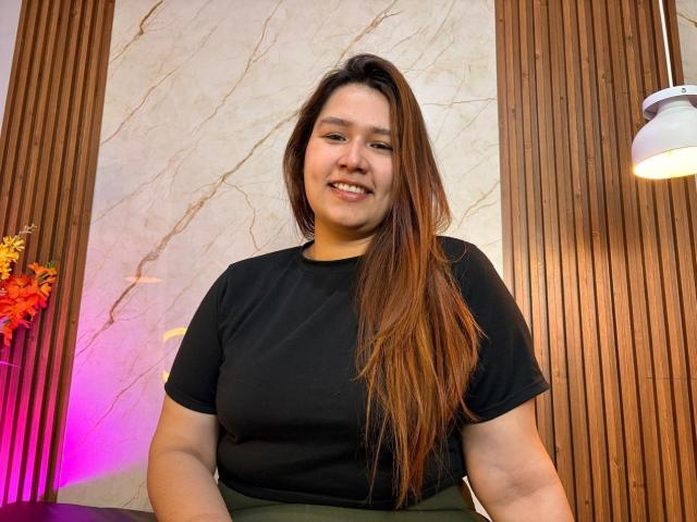 SofiaVelvet, a plus-sized Asian woman with long brown hair, smiling, wearing a black t-shirt, green skirt, and wooden background with a hanging light and pinkish-purple light.