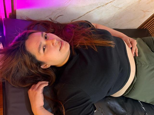 SofiaVelvet lying on a black leather couch, wearing a black shirt and green pants, with long brown hair, looking at the camera, hand on her belly, pink light.