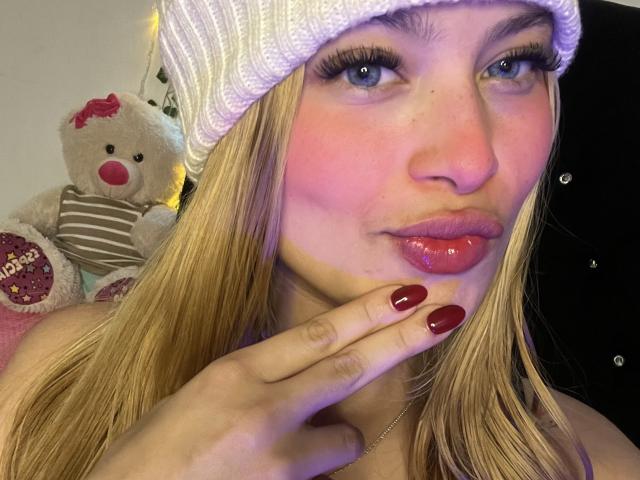 Shantal, con labbra rosse carnose, capelli biondi e un cappello bianco di lana, che indossa uno smalto rosso, toccandosi il mento, con un simpatico orsacchiotto di peluche sullo sfondo..