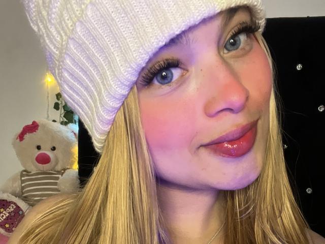 ShantalSummer has blonde hair, blue eyes, and pink lipstick, wearing a white knit beanie. She's smiling with her tongue slightly out, showing off her glossy pink lips.