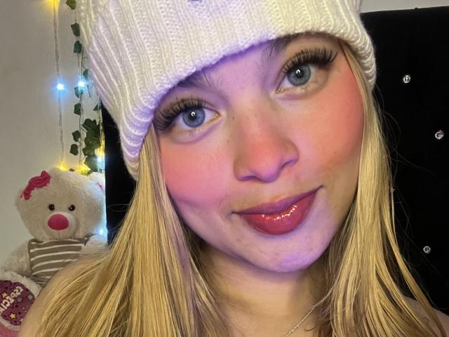 Shantal, im Sommer mit blonden Haaren, einer weißen Strickmütze, rosigem Rouge, glänzenden rosafarbenen Lippen, die Zunge herausgestreckt, langen Wimpern und blauen Augen, hat einen niedlichen, kleinen Teddybären mit rosafarbener Nase im Hintergrund..