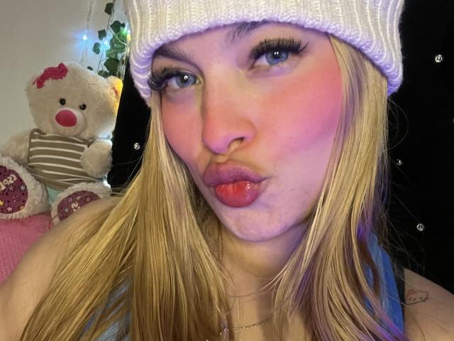 ShantalSummer with pouty lips, blonde hair, white knit beanie, and pink blush, showing off her tongue piercing. Stuffed teddy bear in striped shirt and pink bow in background.