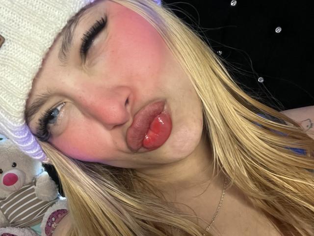 Shantal, im Sommer mit geröteten Wangen, glänzenden roten Lippen und blonden Haaren, die ihre Zunge herausstreckt, trägt eine weiße Strickmütze und hat blaues Augen-Make-up. Ihre Brüste sind leicht sichtbar..
