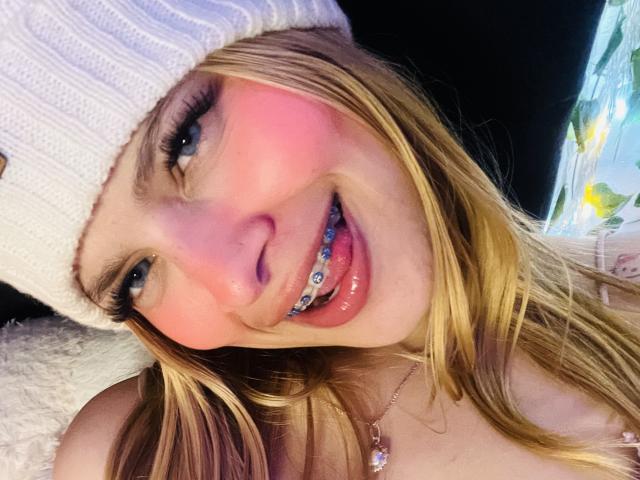 Shantal, con i capelli biondi, le guance rosate, un berretto bianco, denti scintillanti di colore blu e una collana lucida, sorride alla fotocamera. Il suo seno non è visibile..