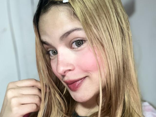 ShantalSummer has blonde hair, pink cheeks, and is smiling with her hand near her face. She has green eyes, light skin, and is wearing pink lipstick.