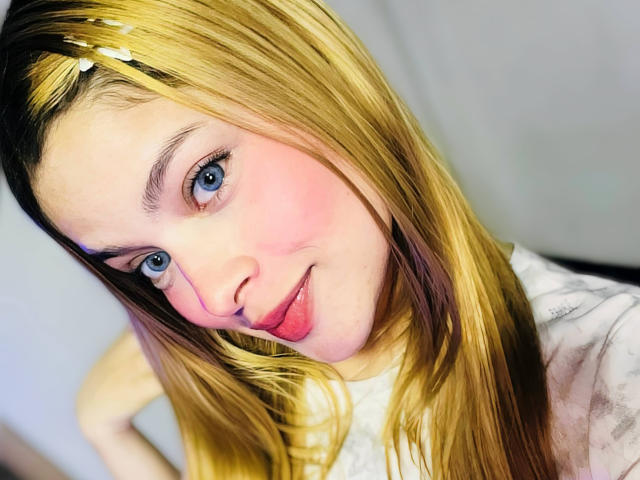 Shantal, im Sommer mit blonden Haaren, rosigen Wangen und blauen Augen, steckt ihre Zunge heraus. Sie trägt ein weißes Oberteil und sieht süß und verspielt aus. Ihre Brüste sind nicht sichtbar..