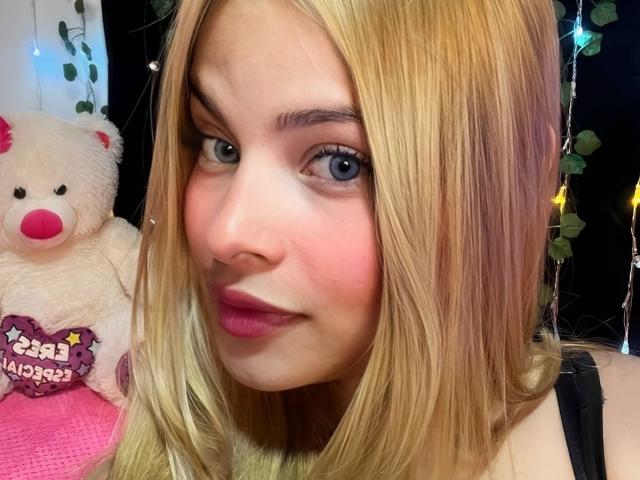 ShantalSummer, com cabelo loiro, batom rosa e olhos azuis, exibindo suas nádegas em um selfie com um adorável ursinho de pelúcia branco. Seu rosto está levemente corado..
