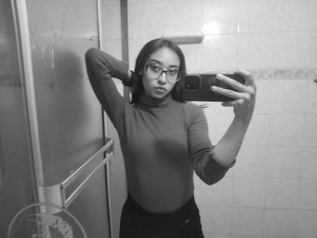 Black-and-white selfie of CamiVargas with glasses, wearing a turtleneck, arms raised, holding a phone, showing a slim, flat-ass, small tits, and dark pants in a tiled bathroom.