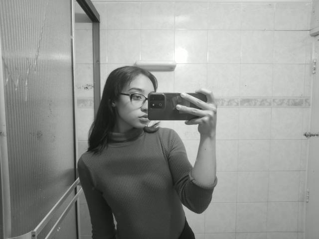 Selfie in bianco e nero di CamiVargas con gli occhiali, che indossa una maglia a maniche lunghe a coste, tiene un telefono, mostra un'espressione accigliata e si trova in un bagno con piastrelle..