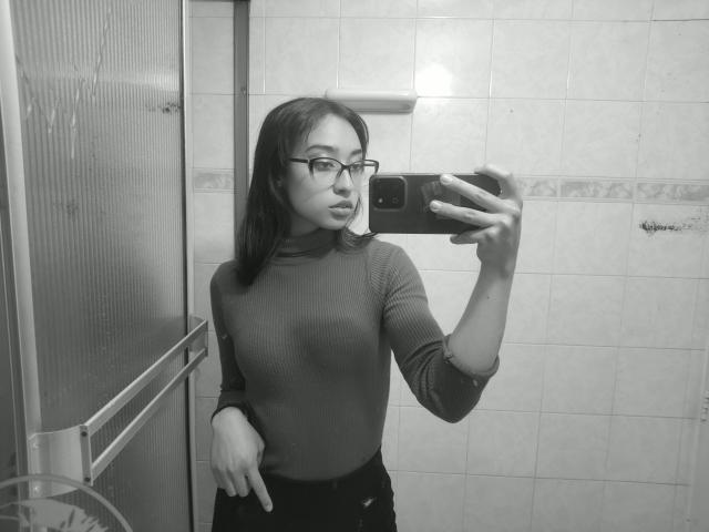 Selfie in bianco e nero di CamiVargas con gli occhiali, capelli scuri di media lunghezza, che indossa un maglione a collo alto a coste, tiene un telefono in un bagno con piastrelle. La sua mano punta verso il basso..