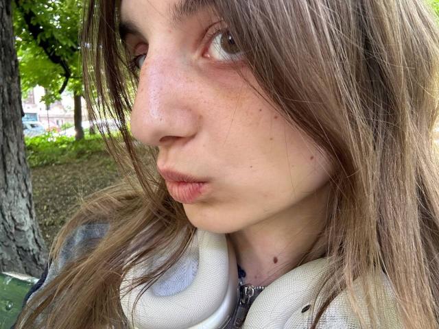 KarieMestlers leicht gerötete, gepolsterte Nase mit kleinen Sommersprossen, braune Augen, die zur Seite schauen, und leicht zusammengepresste Lippen, trägt eine graue Jacke mit weißem Kragen, in einer von Bäumen gesäumten Gegend..