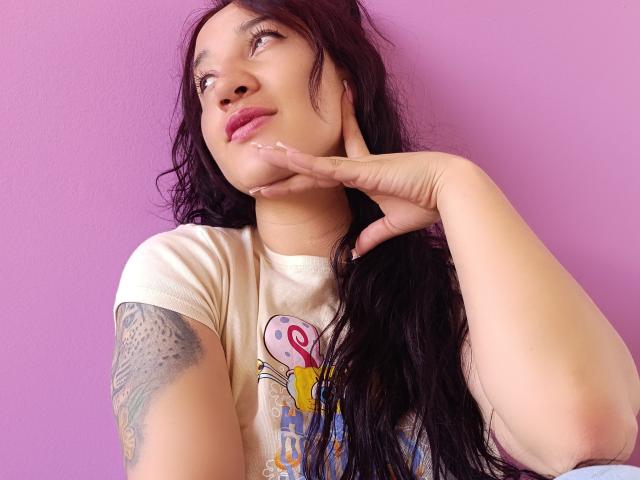 JessiicaDora mit langem, schwarzem Haar, heller Haut und einem Tattoo auf ihrem rechten Arm, posiert vor einer rosa Wand, ihre Hand unter ihrem Kinn, und trägt ein weißes T-Shirt..