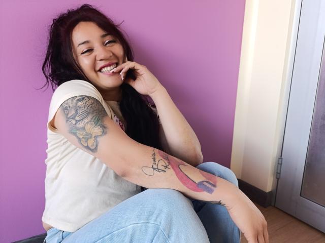 JessiicaDora, con cabello oscuro, sonriendo, camisa blanca, jeans azules, mostrando tatuajes en los brazos, sentada, con un dedo cerca de la boca, pared morada, suelo de madera y puerta de vidrio en el fondo..