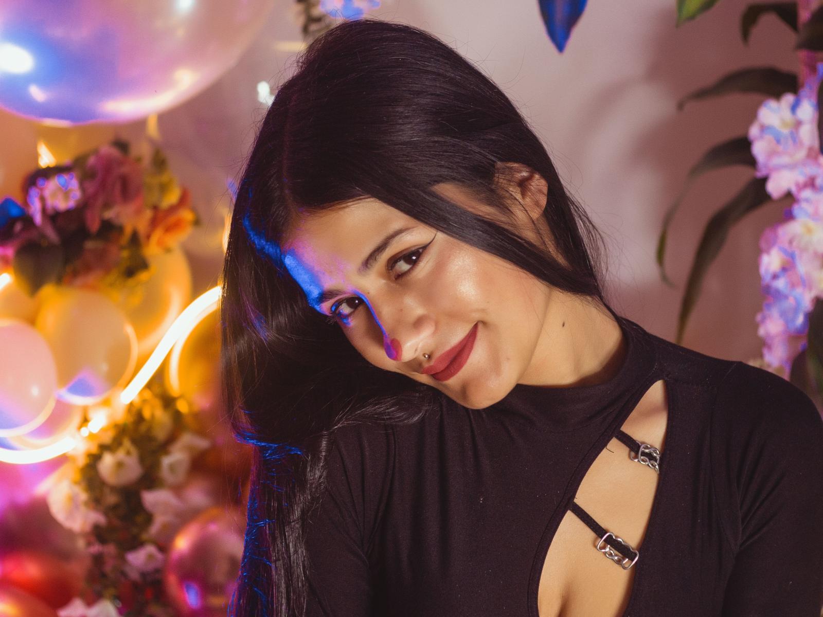 OrianaValens, con el pelo largo y negro, sonriendo, vestida con un vestido negro con tirantes de corte, lápiz labial rosa y un piercing en la nariz, iluminada por luces azules y amarillas..