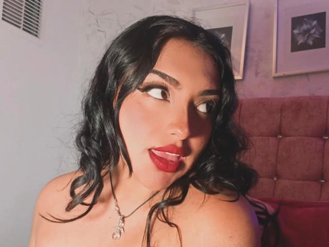 SyraPalmer with black wavy hair, red lipstick, and a necklace, sticking her tongue out, looking to the side, in a bedroom with a pink tufted headboard.