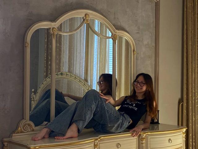 ElmaGalizia, barefoot with glasses, jeans, and black top, lounges on a gold mirror-fronted dresser, reflected in mirror, smiling, long brown hair, slender, small tits, relaxed.