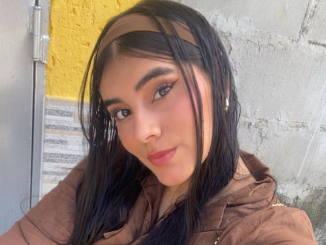 AlaraKey has long black hair, brown headband, brown makeup, and brown hoodie, with a cute smile, showing her nose and lips, and wearing small earrings.