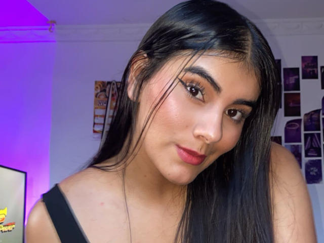 AlaraKey has long black hair, brown eyes, and red lipstick, wearing a black top, with a subtle smile, and a slight nose tilt, showing a cute, sexy look.
