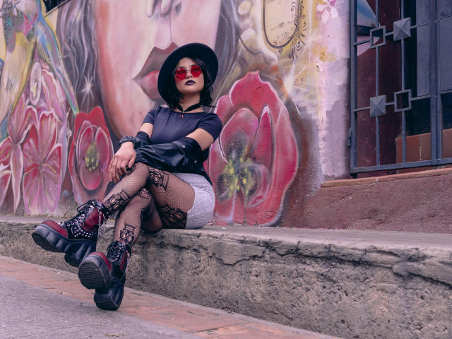 CassieJones with black hat, red sunglasses, black shirt, white shorts, fishnet stockings, tattoos, and chunky boots, sitting on a concrete ledge, showing her ass, in a gritty urban setting.