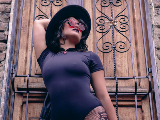 CassieJones with black dress, hat, and red lipstick, standing in front of wooden door with iron scrollwork, wearing fishnet stockings, her hand on her head, showing her tits.