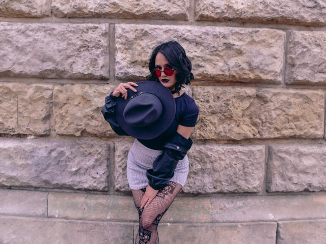 CassieJones with dark lipstick, red sunglasses, black off-shoulder top, grey pinstripe skirt, black hat, fishnet stockings, holding hat against rough stone wall. Curvy, sexy, black wavy hair.