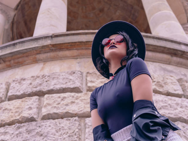 CassieJones has dark skin, wears a black hat, red sunglasses, black top, and black jacket, with a gray waistcoat, showing her tits slightly through the top.